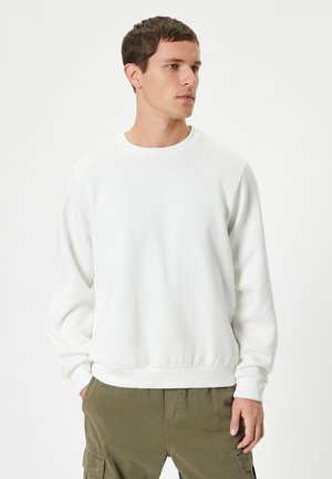 Sweatshirt bianco a collo rotondo realizzato in tessuto morbido, con polsini e orlo a costine. Indossato con pantaloni cargo verde oliva, che mostrano una vestibilità rilassata.