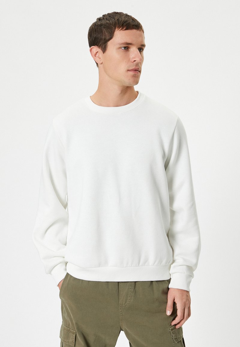 Sweatshirt bianco a collo rotondo realizzato in tessuto morbido, con polsini e orlo a costine. Indossato con pantaloni cargo verde oliva, che mostrano una vestibilità rilassata.