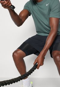 Atletyczny mężczyzna w zielonej koszulce Nike i czarnych szortach trzyma teksturowaną czarną linę treningową, wykonując ćwiczenie.