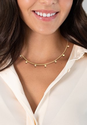 Collier doré avec une chaîne délicate, comportant de petites perles rondes et des variations subtiles dans la taille des perles, porté sur une blouse de couleur claire.