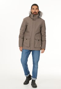 Brun polstret jakke med hætte af falsk pels, lynlåslommer i siderne og lommer med klap og knap, båret med blå jeans og sorte støvler.