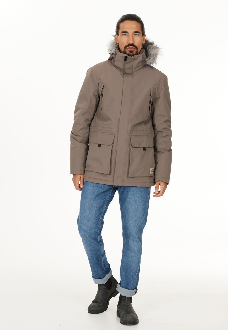 Brun polstret jakke med hætte af falsk pels, lynlåslommer i siderne og lommer med klap og knap, båret med blå jeans og sorte støvler.