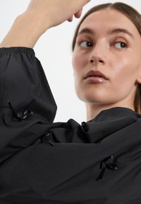 Top cropped nero con colletto alto, maniche lunghe arricciate con polsini elastici e dettagli decorativi in nastro nero. Tessuto liscio.