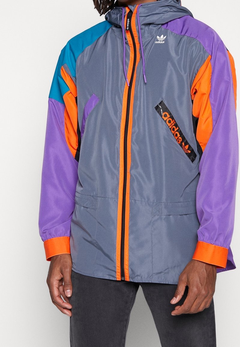 Grå vindjacka med orange, lila och teal-accenter. Har dragkedja fram, två fickor och en Adidas-logga. Slät textur.