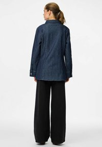Camisa de mezclilla con rayas blancas verticales, puños con botones y un cuello clásico, llevada sobre pantalones anchos negros.