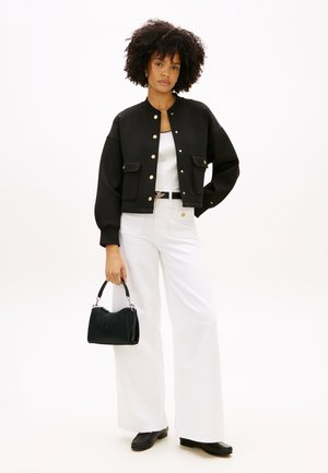 Femme aux cheveux bouclés portant une veste noire boutonnée, un haut blanc et un pantalon large blanc, tenant un petit sac à main noir.