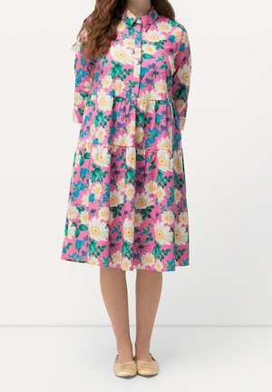 Vrouw draagt een roze midi-jurk met knopen, gelaagd, met wit- en blauw bloemenpatroon, gecombineerd met beige platte schoenen op een witte achtergrond.