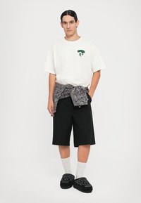 T-shirt de algodão branca com gráfico verde, calções pretos, faixa cinza com padrão na cintura, meias brancas e chinelos pretos com padrão.