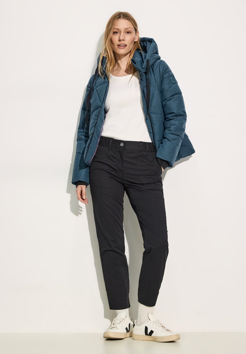 Blauwe gewatteerde pufferjack met een gestikte structuur, witte lange mouwen top, zwarte cargobroek en witte sneakers met zwarte accenten.