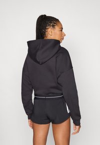 Vrouw met gevlochten haar, gekleed in een zwarte capuchontrui en zwarte sportshorts, opzij gekeken tegen een effen achtergrond.