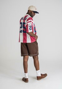 Tricou cu dungi roșii și albe, cu mâneci scurte și accente albastre, având inscriptionat „25” pe spate, asortat cu pantaloni scurți maro și pantofi maro.