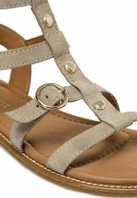 Sandalo in suede beige con più cinturini, hardware decorativo dorato e dettaglio a cerchio in metallo. Soletta imbottita marrone e bordi cuciti.