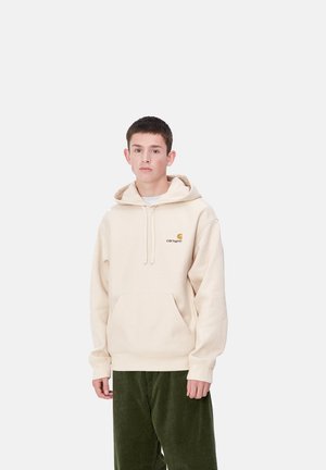 Sudadera beige con bolsillo frontal, capucha con cordones y logo bordado. Combinada con pantalones de pana verdes. Diseño simple y casual.