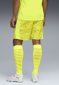 Maglia e pantaloni sportivi giallo neon con motivi grafici, abbinati a calze al ginocchio e scarpe da calcio bianche. Design texturizzato e slanciato.