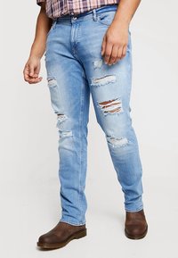 Uomo che indossa jeans strappati di colore azzurro chiaro abbinati a stivali di pelle marrone e una camicia a quadri, in piedi davanti a uno sfondo bianco.