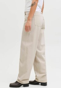 Jean large en denim de coton beige clair, avec une taille haute, des poches arrière et des coutures visibles. Assorti à des bottes noires à talons bas.