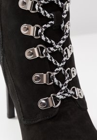 Black suede ankle boot featuring silver hardware and a braided black and white lace design along the front, with a pointed toe and high heel.