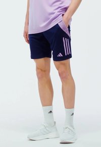 Pantalones cortos deportivos azul marino con acentos en rosa de tres rayas, con bolsillos laterales y una tela flexible y transpirable. Zapatillas y calcetines blancos.