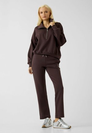 Donna con lunghi capelli biondi indossa felpa marrone scuro con zip e pantaloni coordinati con sneakers argento, in piedi davanti a uno sfondo semplice.