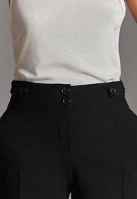 Zwarte hoge taillebroeken met een gladde textuur, voorzien van dubbele knoopsluitingen en plooien aan de voorkant. Gepaard met een witte geribde tanktop.