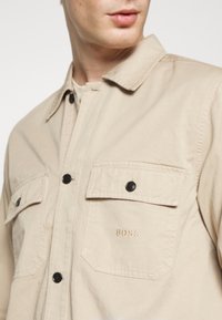 Beige košile s knoflíky vyrobená z bavlny, se dvěma náprsními kapsami, černými knoflíky a vyšitým logem "BOSS" na levé kapse.