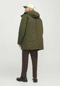 Chaqueta acolchada verde oliva con capucha, tejido texturizado y detalles ajustables, combinada con pantalones marrón oscuro y zapatos negros.