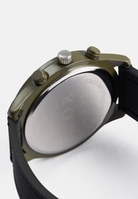 Ελαιοφόρος πράσινος ανοξείδωτος χάλυβας ρολόι με μαύρη υφασμάτινη λωρίδα, διαθέτει δύο κουμπιά και μία κορώνα, ασημένιες λεπτομέρειες και γυαλιστερό μεταλλικό πίσω μέρος.