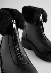 Black leather ankle boots featuring a plush black faux fur cuff, silver zippers, and a chunky black heel. Smooth texture with contrasting stitching.