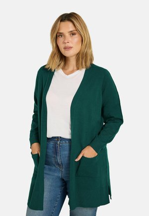 Femme portant un cardigan vert foncé ouvert sur un t-shirt blanc et un jean bleu, debout les mains dans les poches devant un fond uni.