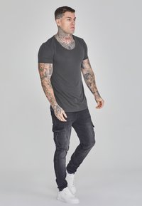 Černé tričko s krátkým rukávem v kombinaci s tmavými cargo kalhotami a bílými vysokými teniskami. Obsahuje podrobné tetování na pažích a krku.