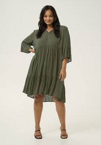 Vestido verde oliva de capas, hecho de una tela ligera, con escote en V, mangas de campana de tres cuartos y un dobladillo desigual.