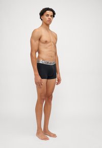 Črne bombažne bokserice z srebrnim pasom z logotipom Calvin Klein, ki nudijo tesen prileganje in gladko teksturo, model stoji bosa.