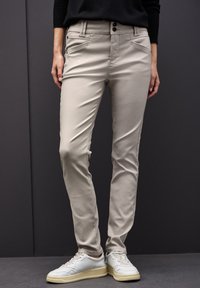 Beżowe spodnie slim-fit o gładkiej fakturze, z dwoma przednimi kieszeniami, zapięciem na guzik i subtelnymi detalami przeszycia. Połączone z białymi trampkami.