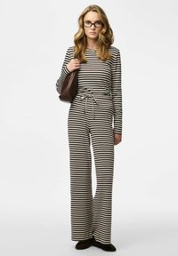 Tuta a righe a maniche lunghe in bianco e nero con vita regolabile a coulisse. La modella tiene in mano una borsa marrone, indossa ballerine in camoscio scuro e occhiali.