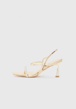 Złoty sandał na cienkich paseczkach z kwadratowym noskiem, cienkim paskiem wokół kostki i niskim obcasem typu bloczek. Z przodu ozdobiony akcentem z kryształków.