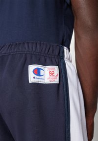 Pantalones cortos deportivos de color azul marino con paneles laterales en blanco y gris oscuro. Presenta un logo bordado y una etiqueta con detalles rojos en la cinturilla.