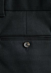 Tissu texturé sombre avec fermeture à bouton. Présente un motif subtil et des détails de couture au-dessus du bouton. Apparence nette et soignée.