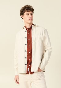 Lichtbeige jas met grijze knopen, twee voorzakken, gedragen over een roestkleurig overhemd met knopen. Glad oppervlak, relaxte pasvorm.