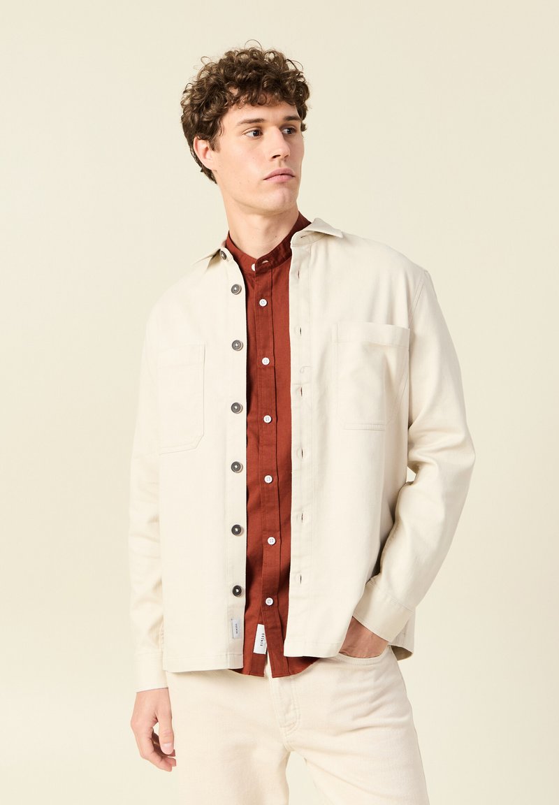 Lichtbeige jas met grijze knopen, twee voorzakken, gedragen over een roestkleurig overhemd met knopen. Glad oppervlak, relaxte pasvorm.