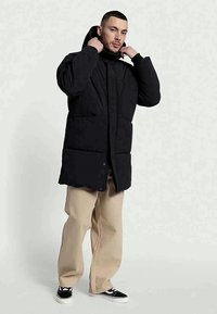 Fekete puffkabát kapucnival, elöl cipzárral, nagy zsebekkel és enyhén hosszú fazonban. Bézs, laza szabású nadrágra húzva.