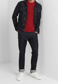 Mand i mørk denimjakke, mørke jeans med opsmøg, rød t-shirt, hvide sneakers, hænderne i lommerne, stående mod en lys ensfarvet væg.