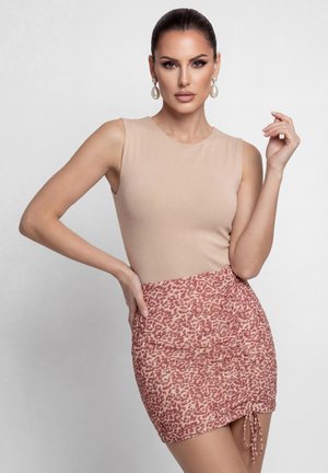 Nainen, jolla on vedetyt hiukset, helmikorvakorut, beige hihaton paita ja vaaleanpunainen leopardi-kuosinen ryppyinen minihame, poseeraa yksiväristä taustaa vasten.