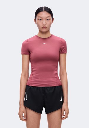 Ung kvinde med sort, glat hår iført en pink tætsiddende kortærmet top og sorte træningsshorts, stående mod en ensfarvet lys baggrund.
