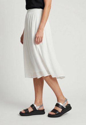 Vrouw draagt een witte midirok en zwarte sandalen met zilveren bandjes, staand op een effen lichte achtergrond.