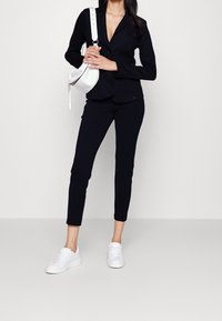 Blazer nero sartoriale con revers, pantaloni neri aderenti abbinati e sneakers bianche. La modella tiene una borsa curva bianca.