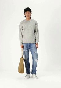 Junger Mann steht, trägt einen grauen Pullover, zerrissene blaue Jeans, weiße Turnschuhe und hält eine beigefarbene Tragetasche an seiner Seite.