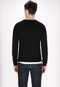 Maglione nero a maniche lunghe con scollatura rotonda, texture liscia e taglio aderente, indossato sopra una camicia bianca, abbinato a jeans in denim scuri.