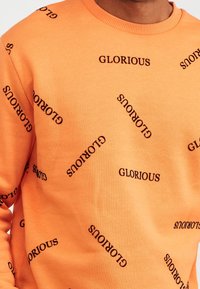 Sudadera naranja con texto negro "GLORIOUS" impreso en varias orientaciones. Cuello redondo acanalado y textura suave.
