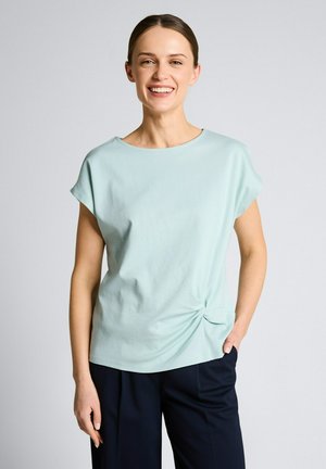 Uśmiechnięta kobieta z brązowymi włosami związanymi do tyłu, ubrana w jasnoniebieski top z krótkim rękawem z węzłem po boku oraz ciemnogranatowe spodnie, stojąca na jednolitym tle.