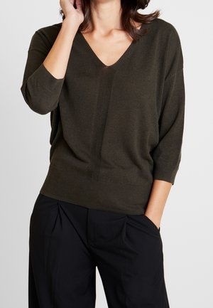 Kvinde iført en mørkegrøn V-hals sweater med kvartlange ærmer og sorte plisserede bukser, den ene hånd rører ansigtet, den anden er i lommen.